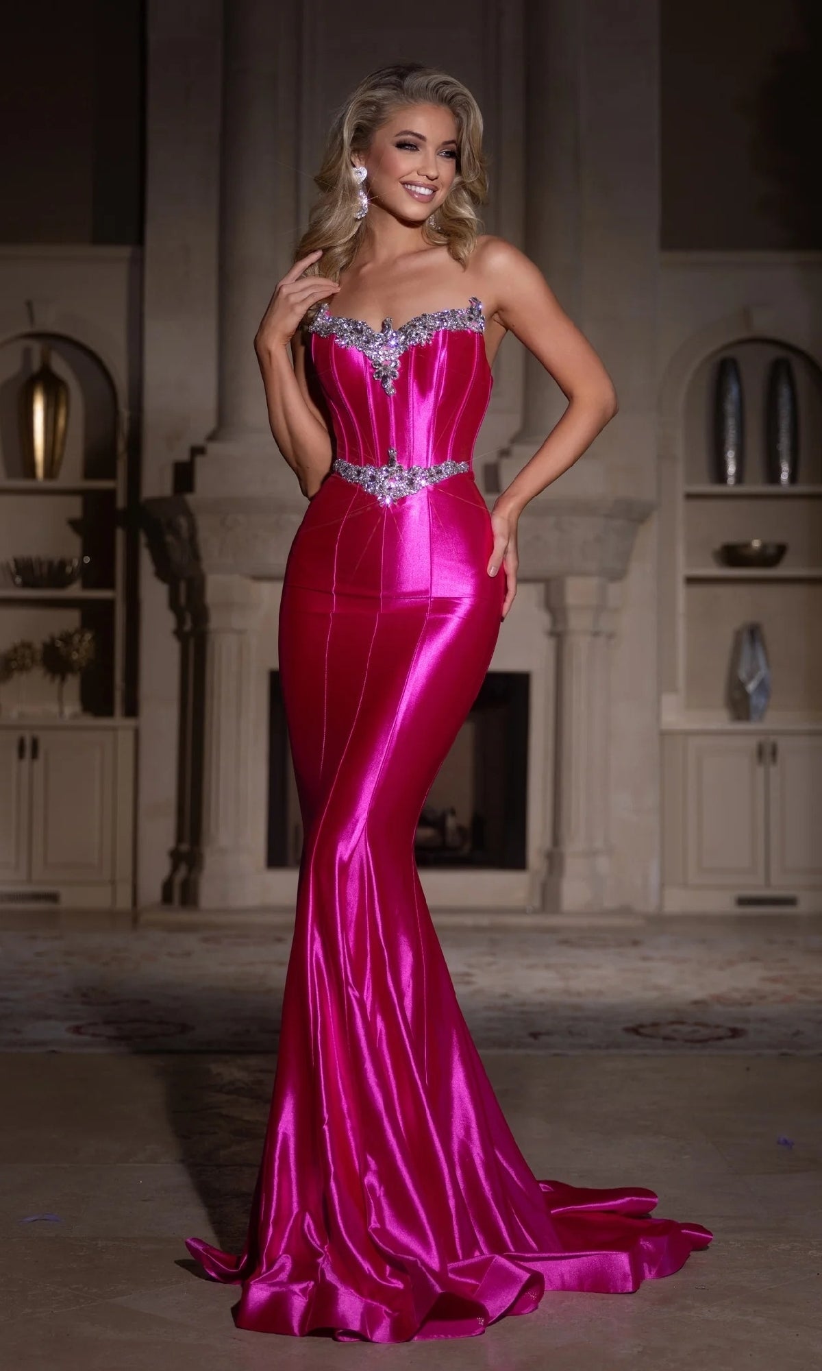 A woman stands indoors wearing the Portia & Scarlett PS25261 long mermaid prom dress in hot pink, featuring fitted strapless satin with jeweled details at the neckline and waist.